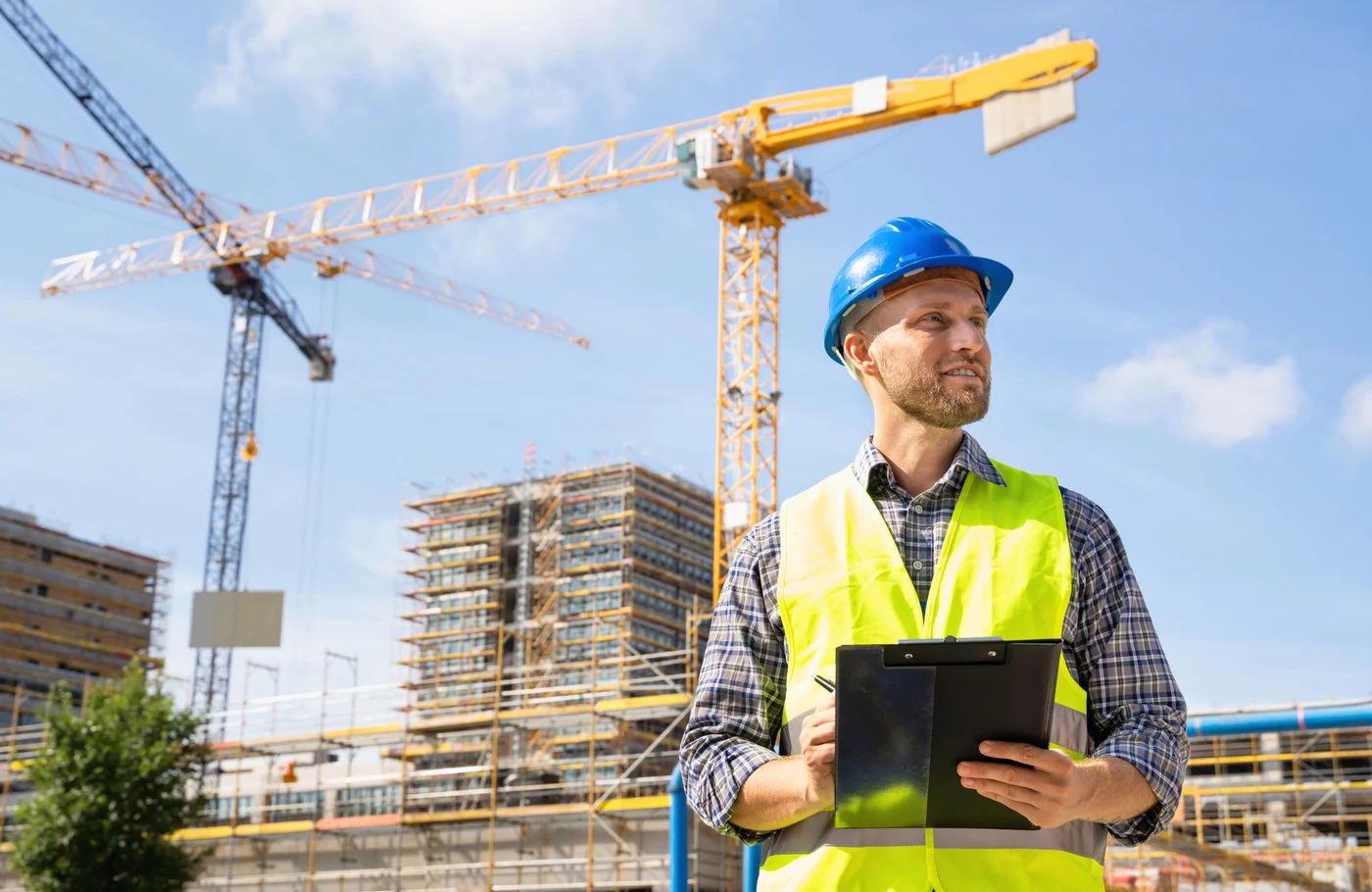 Chargé de projet sur un chantier, planchette à pince en main, devant des grues et un bâtiment en construction